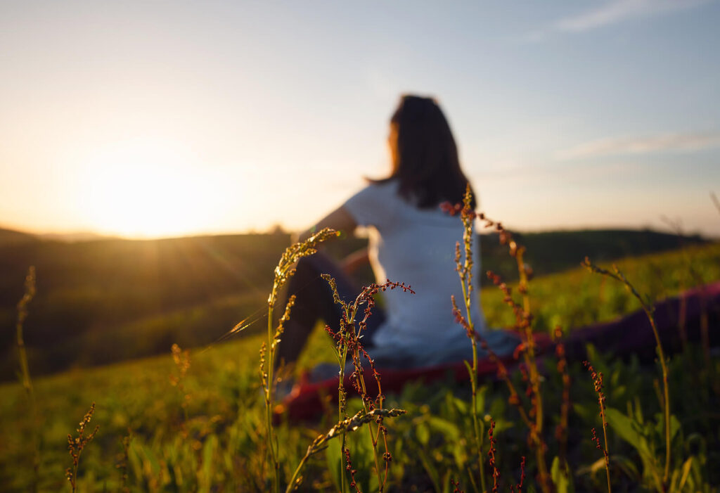 Woman sitting on a hillside watching the sunset. Mindfulness-based therapy in Virginia can help with anxiety, depression, trauma and more. Reach out for mindfulness therapy via online therapy in Virginia.