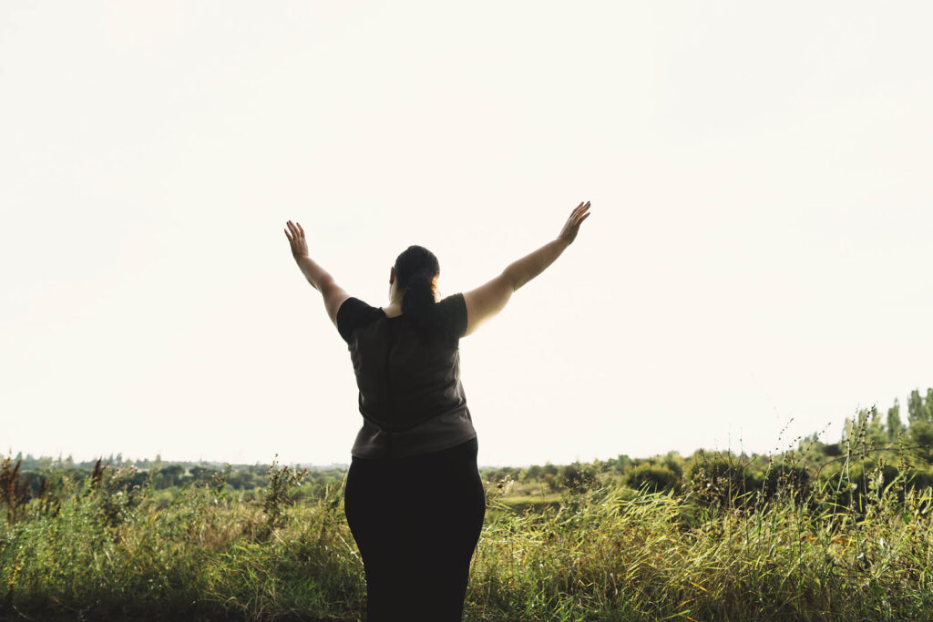 A woman holds her arms up while looking out over a field. This could symbolize the self confidence gained by therapy for self esteem in Virginia. Learn more about therapy for anxiety and other services by contacting an equine sports therapist in Richmond, VA today. 