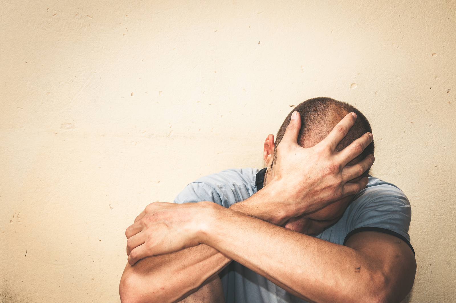 A man covers his head while sitting against a blank wall. This could represent the pain of past trauma that emdr therapy in Richmond, VA can help you address. Learn how trauma therapy in Richmond, VA can help by contacting a therapist for men in powhatan, va
