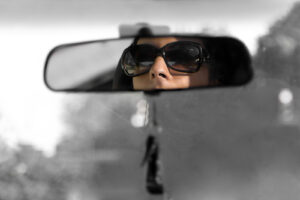 A woman wearing glasses looks in a rear view mirror while driving, recognizing traits inherited from their parents. Learn more about how a narcissism therapist in Richmond, VA can offer support in coping with narcissistic parents. Search for online therapy in Virginia or search for narcissism therapy in Powhatan, VA for support. 