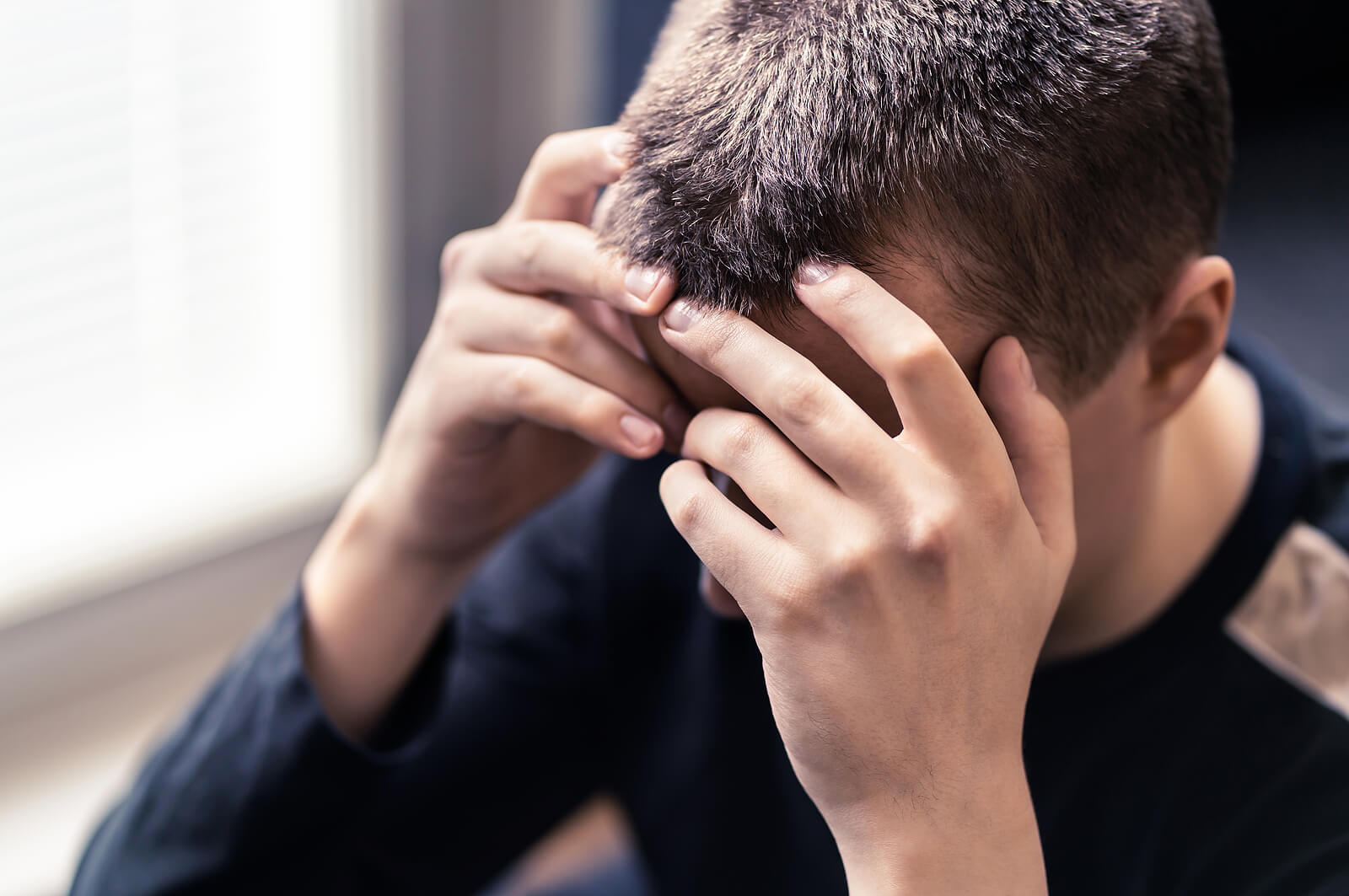 A young man with short hair sits with his head in his hands, looking down in distress. His fingers are interlaced over his forehead. This could represent the stress of hidden trauma that men can face. Learn more about the help therapy for men in Powhatan, VA can help. Search for trauma therapy in Richmond, VA today.