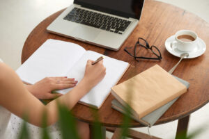 A person writing in a journal at a wooden table with a laptop, glasses, a cup of coffee, and books nearby. This could symbolize journaling to help cope with past narcissism. Learn how narcissism therapy in Powhatan, VA can offer support by contacting a trauma therapist in Richmond, VA. 