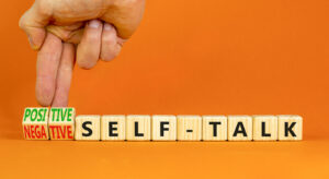 Wooden blocks spelling "SELF-TALK" are displayed on an orange background. A hand is flipping a small wooden block from "NEGATIVE" to "POSITIVE," symbolizing the shift from negative to positive self-talk. Contact an equestrian therapist new kent VA to learn more about how equine sports therapy in Virginia and how equine assisted therapy in Richmond, VA can support you today. 