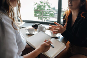 A woman gestures while talking with another person holding a notebook during a supportive conversation over coffee. This scene reflects the connection and trust built with an EMDR therapist in Powhatan, VA, the collaborative process of trauma therapy in Richmond, VA, and the guidance offered by an experienced EMDR therapist in Richmond, VA. 