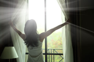 A woman throws open wide curtains to reveal a flood of warm light streaming through a large window, her posture open and expansive as she faces the brightness ahead. This powerful image captures the shift that many clients describe after completing EMDR intensives near me — when memories that once felt like open wounds finally begin to feel like the past. For those who have worked with an EMDR therapist in Powhatan, VA, this moment of opening toward light and freedom is not just a metaphor — it is the destination the work has always been pointing toward. 