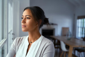 A woman stands with her arms crossed, gazing out a window with a look of quiet contemplation and emotional weight. This kind of inward struggle is something a life transition therapist in Powhatan, VA often sees in clients processing unresolved pain during major life changes. Life transitions therapy in Virginia offers a compassionate space to work through exactly these moments. 
